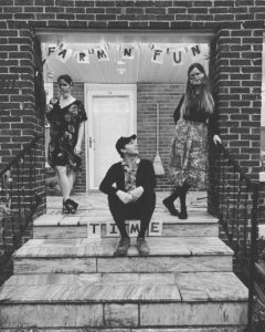 B&W image of Bill & the Belles sitting on a front porch. Kalia and Helen wear face masks with Kris sitting on the stoop below them and the words Farm n' Fun Time hang on a banner above them.