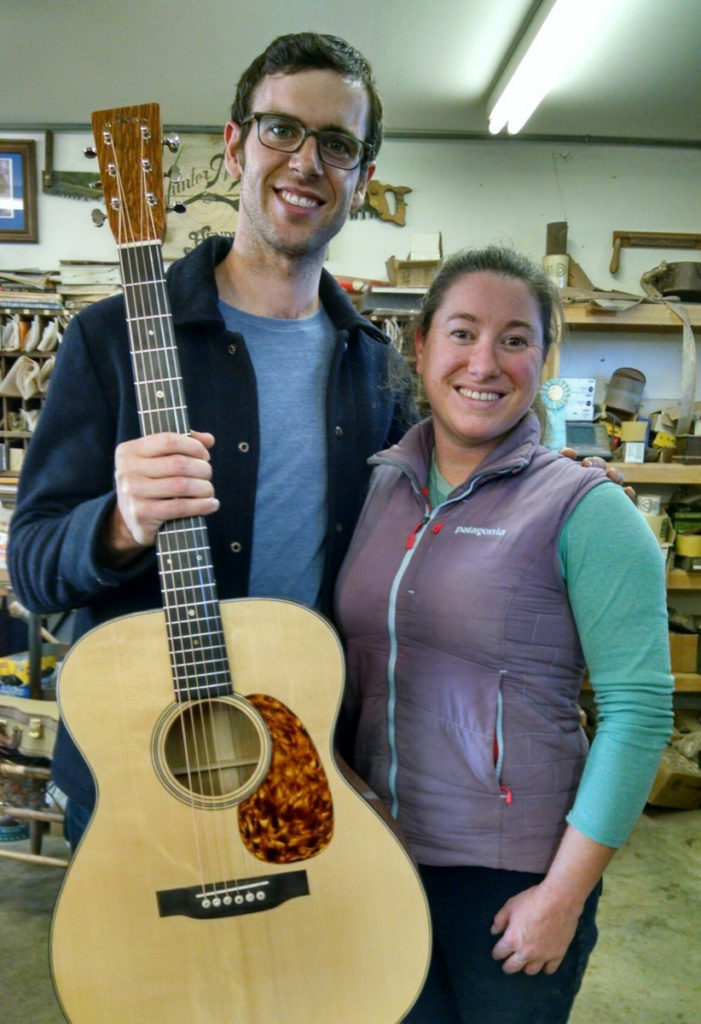 A tall man with Jayne Henderson in a workshop; the man holds the Doc Watson guitar, which is a pale wood with tortoise-like pick guard.