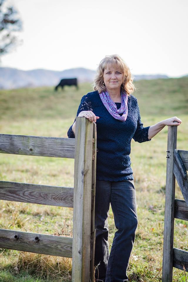 A blonde woman wearing a blue top and pants with a lavender scarf around her neck. She is standing at a wooden fence gate and you can see a black cow behind her in the distance.