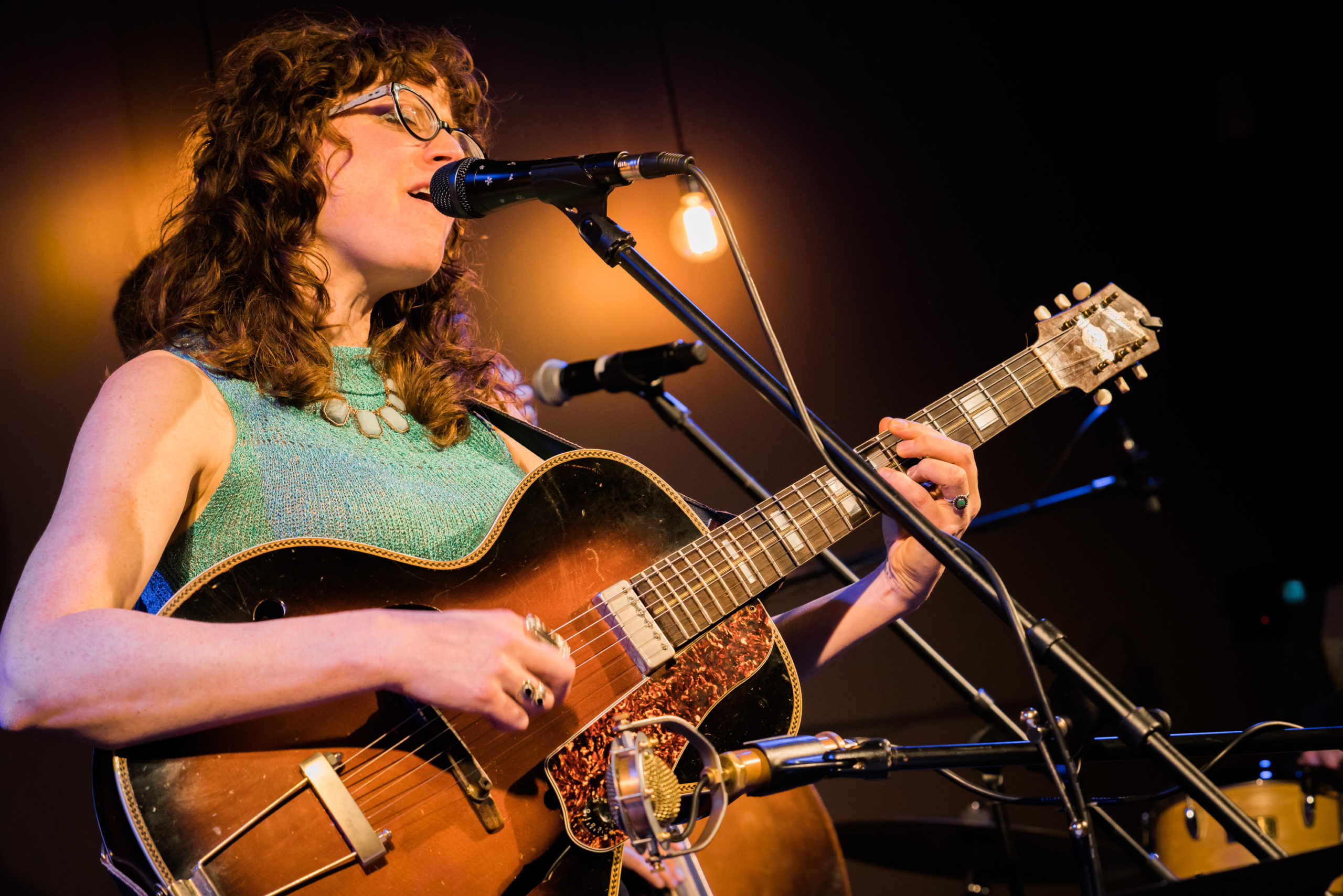 Female guitar player singing at the mic. She has medium-length brown curly hair, glasses, and is wearing a sea green sleeveless top and a white costume jewelry necklace.