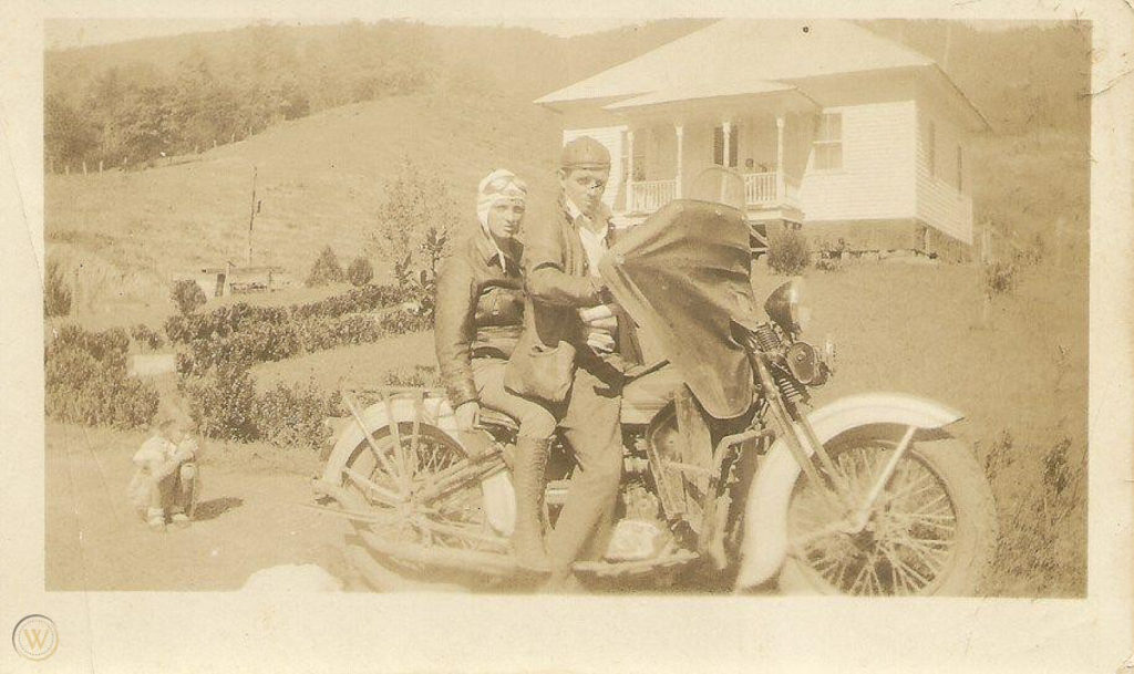 Sepia-style image showing Eck at the front of the motorcycle with Maybelle seated behind him. Both wear helmets. They seem to be in a field or yard in front of a white house.