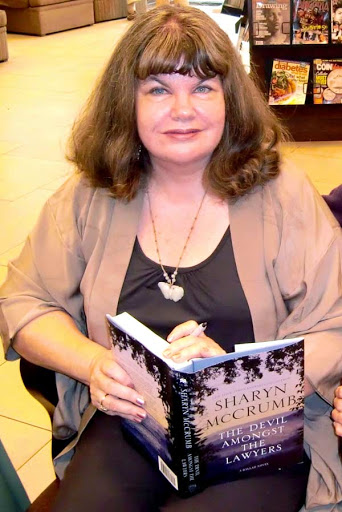 A white woman with shoulder-length brown hair and bangs. She is wearing black pants and a black shirt with a beige cardigan. She is looking at the camera and holding an open book "The Devil Amongst the Lawyers" and a pen, as if she is about to sign the book.