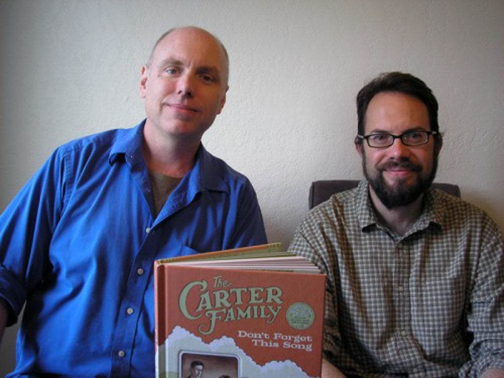 Two white men sit beside each other with the book The Carter Family: Don't Forget This Song in front of them. The man on the left is balding and wears a blue button-down shirt over a grey t-shirt. The man on the right has dark hair and a beard and is wearing a brown-checked button-down shirt. He also has glasses.