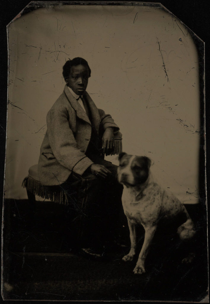 A young Black boy wearing a large light-colored jacket and dark pants sits on a velvet chair or ottoman. A dog that resembles a Staffordshire or pit bull terrier sits at his feet.