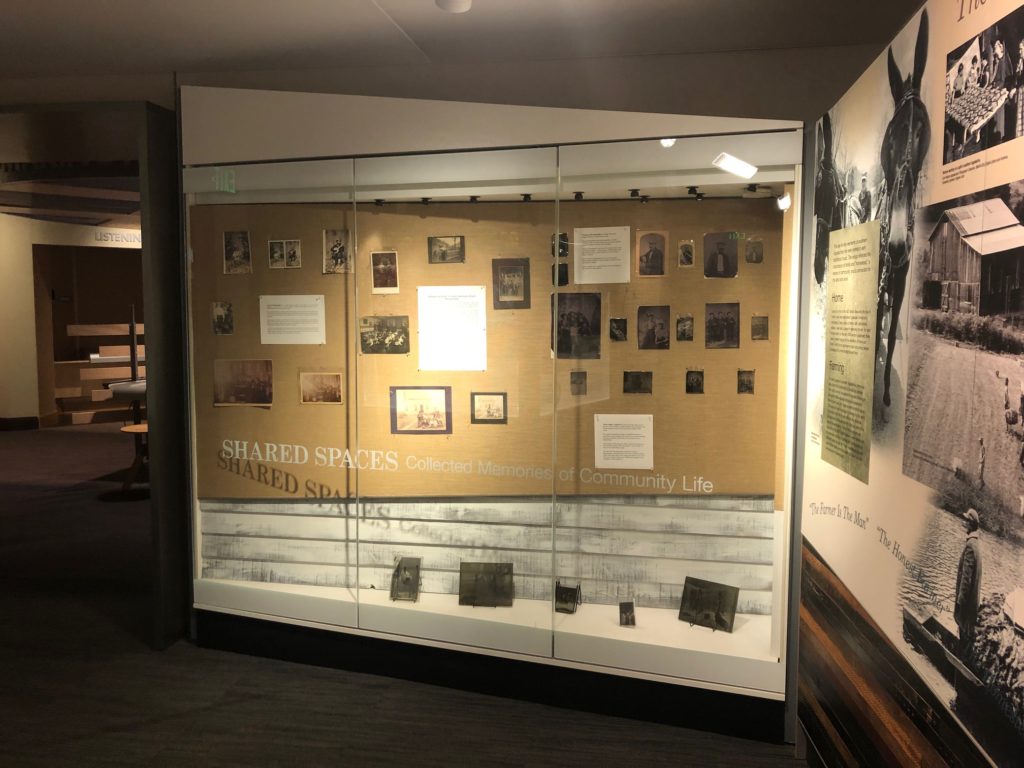 Image shows the Community Case where several photographs are displayed across the back wall of the case, along with explanatory text. Four original glass-plate negatives are arranged in the base of the case, along with a small tintype.