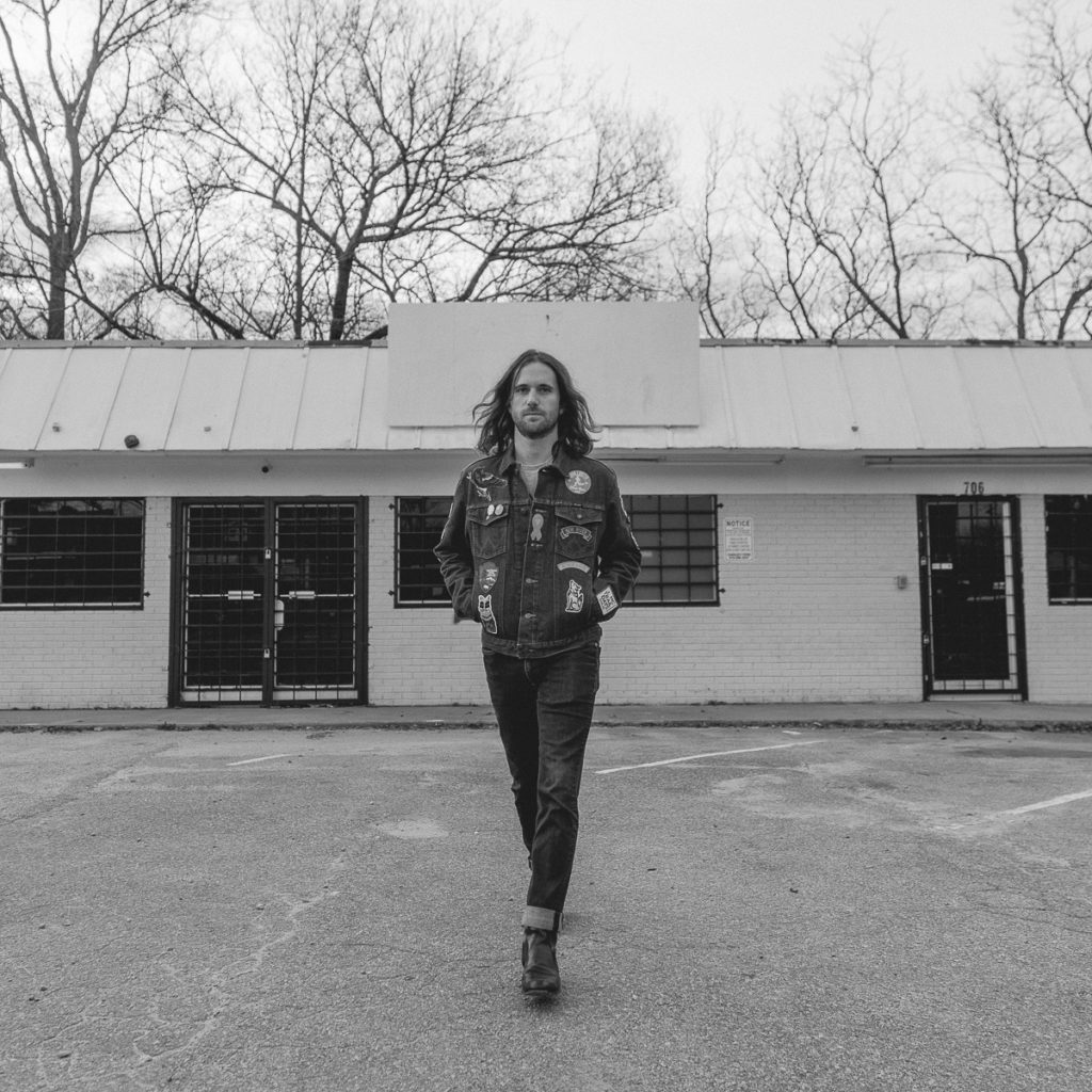 Black-and-white image of a man walking towards the camerain front of what looks to be a closed-down strip mall or old motel. The man has shoulder-length dark hair and a scruffy beard. He is wearing jeans, black shoes, and a jean jacket with numerous patches and badges on it.