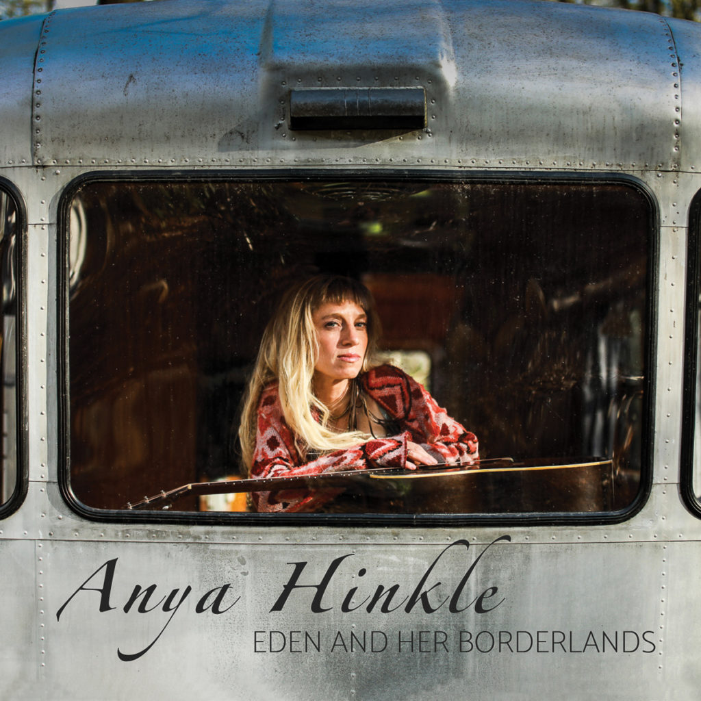 This image is focused in on the back window of a shiny silver Airstream trailer. Anya Hinkle sits in the back window, looking out at the photographer -- she is a blonde white woman, with long hair and waring a red and pink patterned top. She is leaning her arms on a guitar.