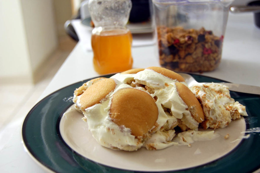 A plate of banana pudding -- showing several Vanilla Wafers on top -- with a plastic bear bottle of honey and a container of granola in the background.