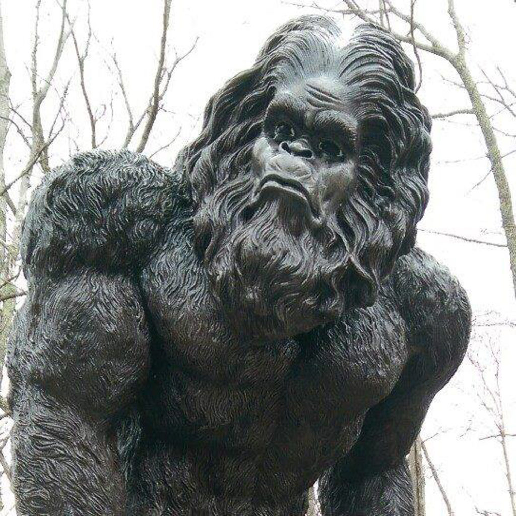A bronze-looking statue of a Bigfoot/yeti-like creature, shown here from the chest up. He is muscular with a furry body, and lots of hair and a beard on his head and face. You can see trees behind him.