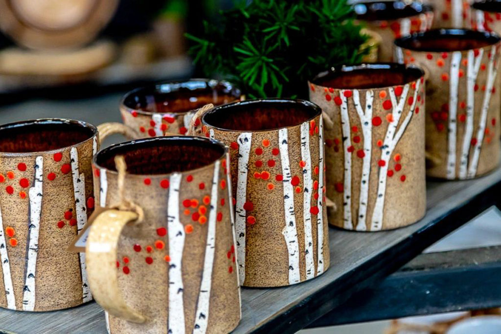 A display of six pottery mugs on a table with green foliage seen behind them. The mugs are colored with a light brown glaze and are decorated with white birch tree trunks and red leaves/berries. The inside of the mugs is a deeper reddish brown.