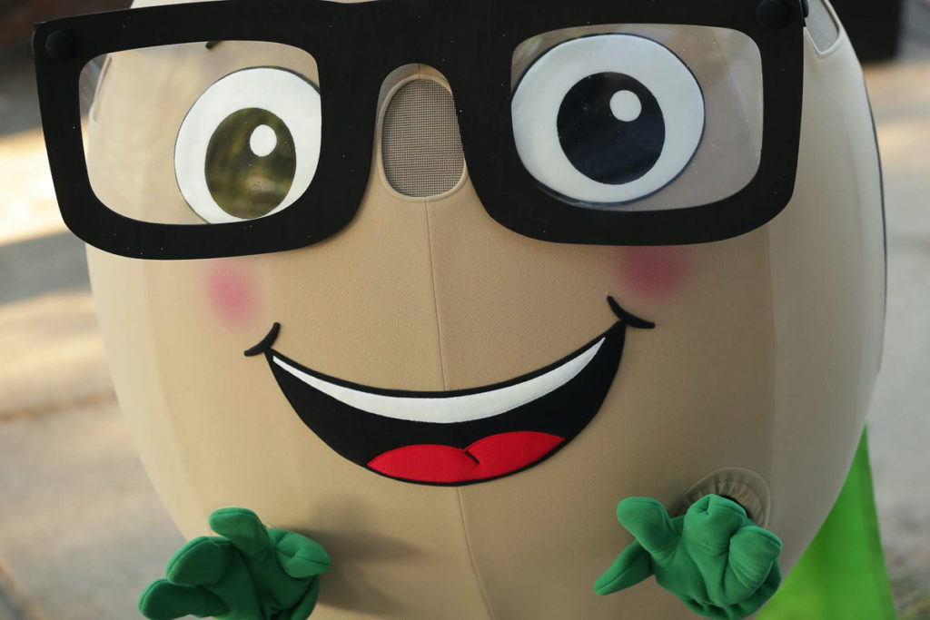 A soybean mascot looks at the camera -- it has a round cream-colored head with black-framed glasses, a big smiling mouth, and two green hands.