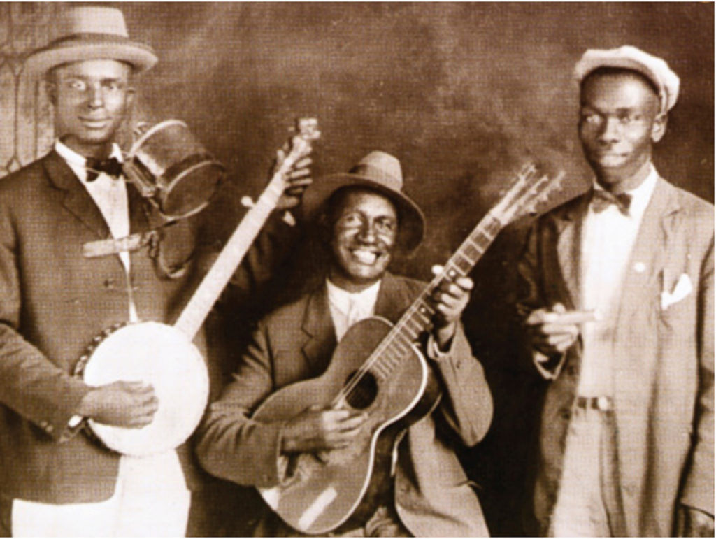 Three Black musicians pose with their instruments. They all wear suits, the men to the left and right with bow ties and the middle man with either a normal tie or just his collar buttoned up. The man to the left wears a fedora-style hat, holds a banjo, and has a jug held towards his face by a "rack" around his neck. The middle man is seated and wears a fedora-style hat; he holds a guitar. The man to the right wears a flat cap and holds a harmonica.