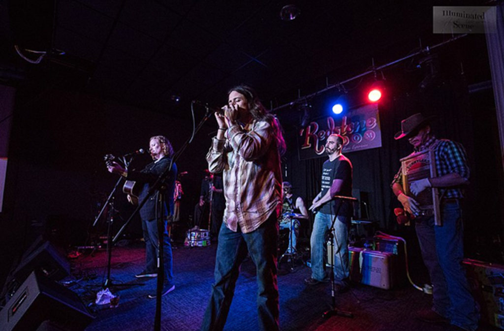 This photograph shows a band on a dark stage. The group is made up of six white musicians, including a woman with curly hair on guitar, a man with longish dark hair and a plaid shirt on harmonica, a man with a black shirt on washtub bass, a man in a tank top on drums, a man in a black shirt with the spoons, and a man wearing a hat and a plaid shirt on the washboard.