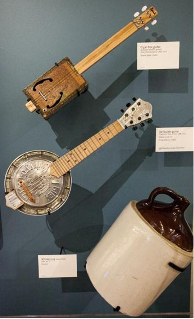 Three instruments hang on the back of a museum display case. The backing is blue. The top instrument is a small guitar made from a cigar box with a wooden neck. The middle instrument is a guitar made out of a circular ice bucket with a wooden neck. The bottom instrument is a large jug with a brown neck and shoulders and a cream body.