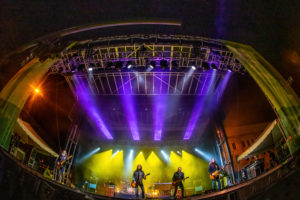 Purple and yellow stage lights shining down on members of the band Blackberry Smoke performing on the State Street Stage at Bristol Rhythm & Roots Reunion 2021. The image has been taken with forced perspective so it seems to curve at the bottom and show the whole stage set-up.