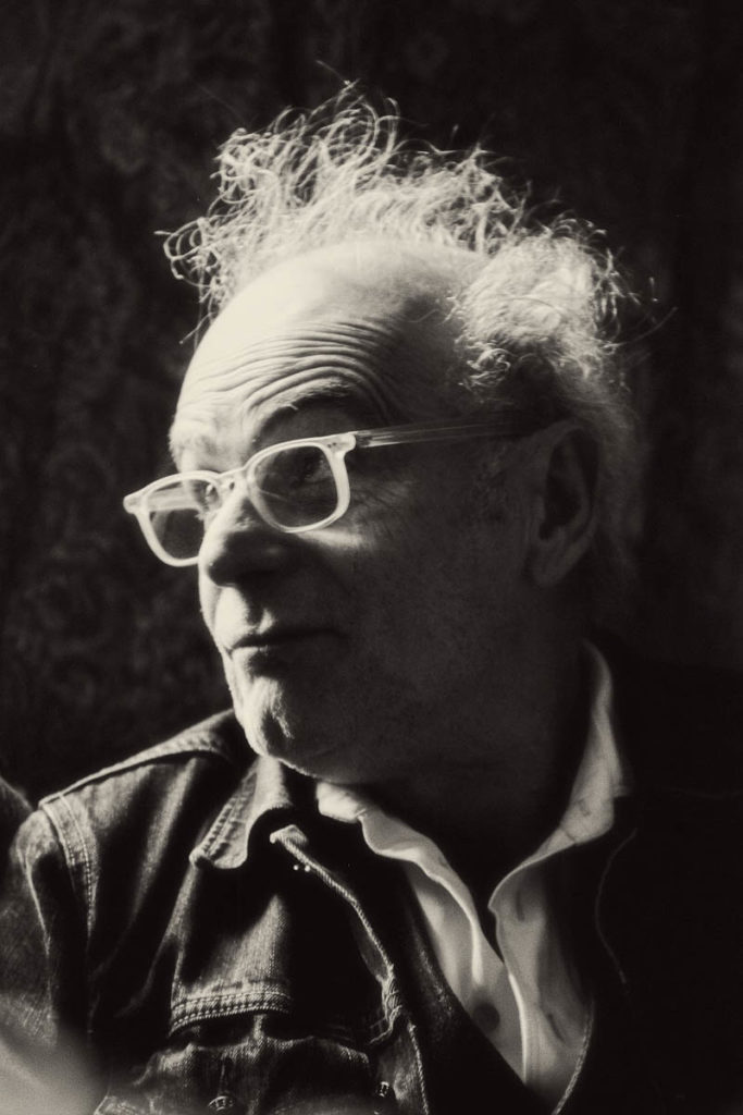 Black-and-white photograph of Ed Snodderly. He is an older white man who wears glasses, a white button-down shirt, and a jean jacket. He is looking up and off to his right. His hair is quite messy and fluffy.