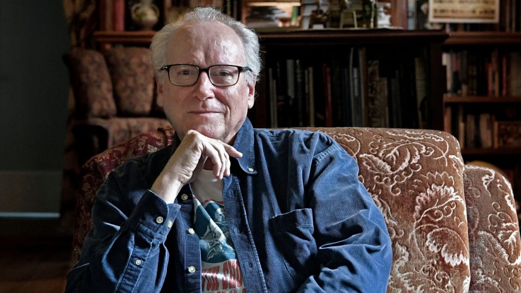 This photograph shows an older white man wearing a dark flue denim button-down shirt over a tee. He has black-rimmed glasses and white thinning hair. He is sitting in a burgandy/brown patterned chair with bookshelves/record shelves and other home decor behind him.