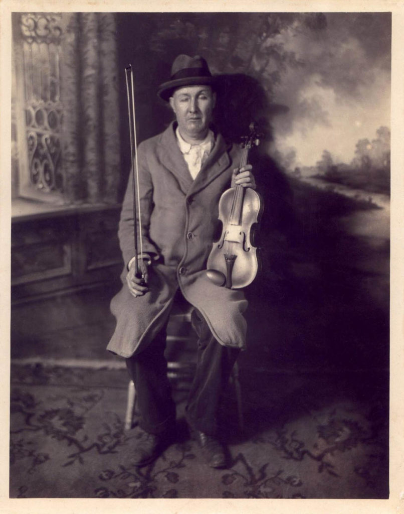 This black-and-white image shows Ed Haley sitting in what looks to be a formalized portrait setting for a photographer. There is a painted landscape behind him and a patterned rug at his feet. He is a white man with partly closed eyes; he wears a dark fedora, a white button-down shirt, dark pants, and a lighter overcoat, buttoned-up. He is holding a fiddle and bow in his hands.