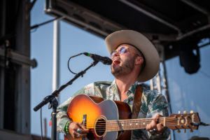 This image shows an African American man playing guitar and singing into a mic on an outdoor stage. He wears a light-colored cowboy hat, sunglasses, and a patterned shirt in blues and greens.