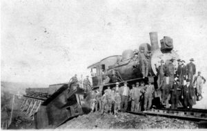 A black-and-white image of a steam locomotive on the railway with the cars behind it derailed from a bridge behind it. Several male workers and bystanders pose in front of the locomotive.
