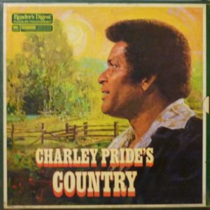 This album cover shows an image of Black musician Charley Pride looking off into the distance over green fenced fields and trees. He is wearing a black jacket with a light-colored collared shirt underneath.