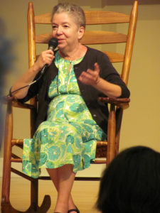 An older white woman sitting in a wooden rocking chair. She is wearing a green-patterned floral dress with a brownish cardigan, and she is holding a microphone while she talks to an audience.
