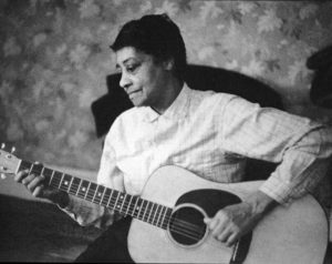 Black-and-white photograph of Black musician Elizabeth Cotten. She is wearing a light-colored long-sleeve shirt and plays a guitar.