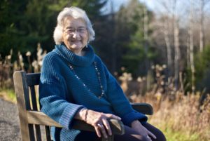 In this image, an elderly white woman sits in a wooden chair with trees and meadow behind her. She is wearing a blue long-sleeved top with a long necklace.