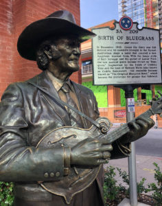 Image of bronze statue of Bill Monroe wearing a suit and cowboy hat and playing his archtop mandolin. A metal historic marker is seen behind Monroe, commemorating the "birth of bluegrass."
