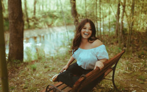 A photo of Emily Scott Robinson seated on a bench in the woods by a creek
