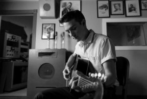 Black-and-white image shows a young dark-haired white man wearing a white shirt and dark pants. He is seated and playing a guitar, and there are various framed images on the wall behind him, along with a display of microphones, and a studio space with various pieces of equipment is seen through a doorway.