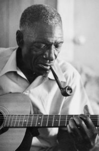 A black-and-white photograph of an older African American man. His hair is cut close to his head, and he wears a white button-down shirt. He is holding a pipe in his mouth and playing a guitar.