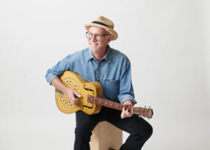 Photograph of an older white man holding a guitar. He is seated on a stool or pedastel, and he wears black pants or jeans, a long-sleeve blue button-down shirt, and a pale straw hat with black band. He also wears glasses.