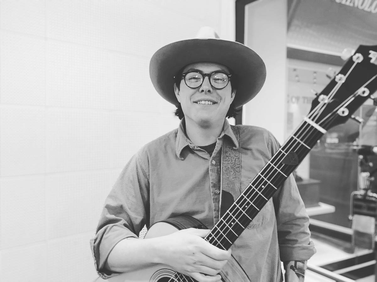 A black-and-white photograph of a white male musician wearing a white Stetson-style hat and holding a guitar. He is standing in front of a radio booth.