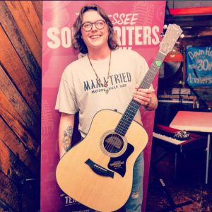 A photograph of a white man standing in front of a vinyl banner for the Tennessee Songwriters Week Competition; you can see a stage area with instruments behind the banner and the man. The man is young, with wavy brown chin-length hair; he is wearing glasses, a white t-shirt, and jeans. He holds a guitar in one hand.