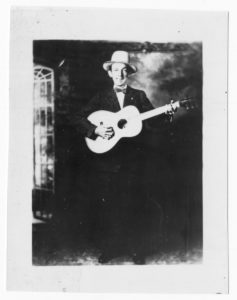 Black-and-white photograph of Jimmie Rodgers. He is a white man, and he stands in front of a faux photographic background that looks like a garden trellis. He is wearing a dark suit, a bow tie, and a white stetson-style hat, and he is holding his guitar.
