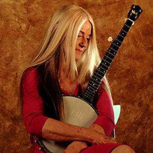 A white woman with long straight white hair wearing a reddish-orange quarter-sleeve dress and a brown vest. She is holding a large head banjo and looking.