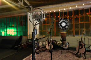 A converted warehouse-style studio space is seen in this photograph. Large multi-paned windows line the back wall; it is night-time outside and you can see the glow of greenish lights in the windows to the left. In the studio can be seen various vintage microphones, instruments on stands, and other music paraphernalia. A record-shaped sign for American Vinyl Co. is hanging in the windows to the right.