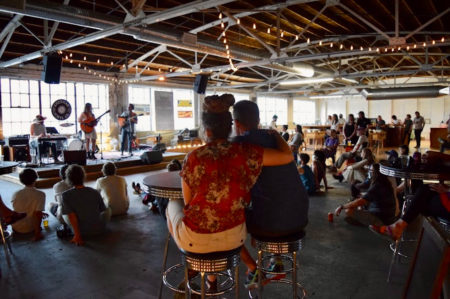 A large warehouse space with a band (a keyboardist and two guitarists) on stage in front of multi-paned windows and a crowd on listeners sitting on the floor and stools in front of them.