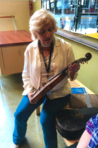 An older white woman is sitting on a wooden bench in a museum space. She has short blond/white hair, and she is wearing a white long-sleeved top over a light-colored tee and dark blue pants. She is holding a fiddle in two hands.