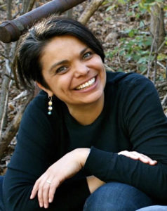 A photograph of a Native American woman. She has short black hair and is wearing a dark blue long-sleeved top and dangly earrings. She is smiling at the camera.