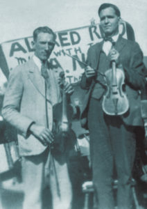 Black and white photograph of two musicians standing in front of a handwritten performance advertiseman placard. Both are white mean and wearing suits and holding fiddles. Blind Alfred Reed is to the right -- he is tall with dark hair. The man to the right is shorter with lighter colored hair.