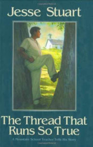 Cover image of The Thread That Runs So True has an illustration of a young white man wearing light-colored shirt and pants leaning against a tree with a one-room white clapboard schoolhouse in the background.