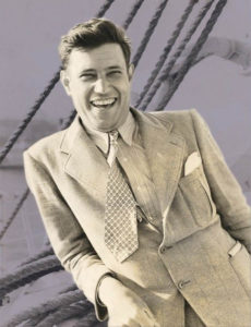 A colorized photograph of Jesse Stuart. He is a white man with dark hair, and he wears a light-colored suit and tie. It looks like he is leaning on the side of a boat in this photograph. He is smiling widely at the camera.