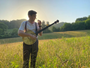 A white man holding a banjo in a sunny field with the hills and trees rising behind him. He holds a flower up to his face to smell it.He wears dark trousers, a white shirt, and suspenders. His hair is brown and cut short.