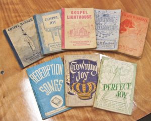 Eight paperback songbooks lie on a wooden table. The books have differently colored covers and a variety of titles.