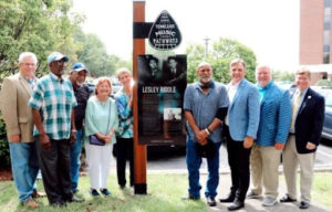 Photograph of nine Black and white men and women standing around the TN Music Pathways marker dedicated to Lesley Riddle. The marker includes a panel with information and a photograph of Lesley Riddle with the guitar pick-shaped TN Music Pathways icon above it.