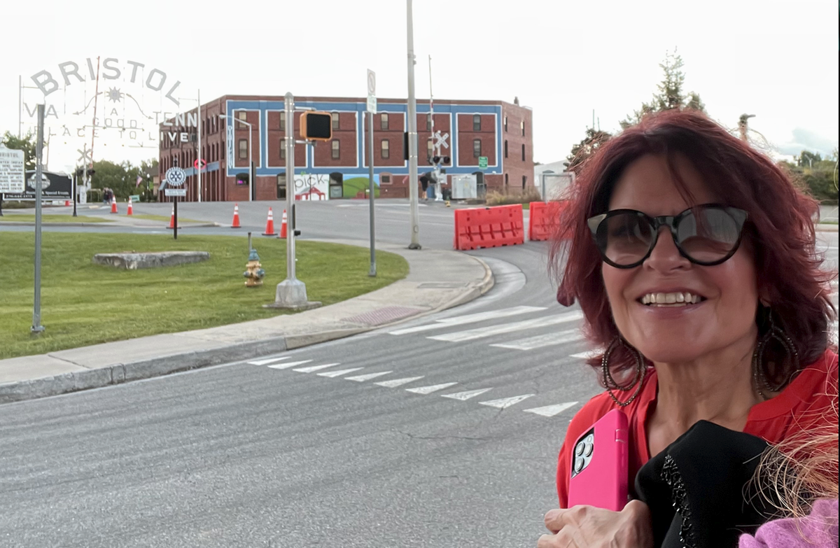 Windblown Rosanne Cash, smiling and wearing sunglass, as she passes the famous Bristol sign.