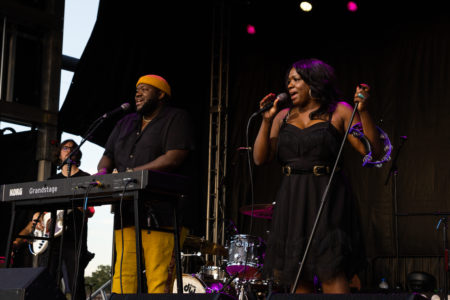 Two black musicians on stage. The man is to the left and is playing keyboards; he is wearing mustard-colored pants, a black short-sleeved shirt, and an orange beanie. He has a beard. The woman is to the right and she is singing into a mic. She is wearing a black belted and thin-strapped dress with fringe along the top of the bodice; she has gold hoop earrings on and has a purple tambourine over one arm. Her hair is shoulder-length.