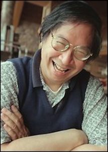 A Chinese American man with dark, slightly graying, hair is smiling at the camera. He wears glasses, a greenish-blue checked shirt with a dark blue fleece or sweater vest over it. He has his arms crossed. 